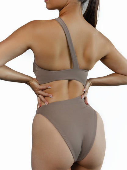 A close-up of a model showing the back side of the Brooklyn one-piece cut-out swimsuit in Truffle color. Model is standing in front of white wall.